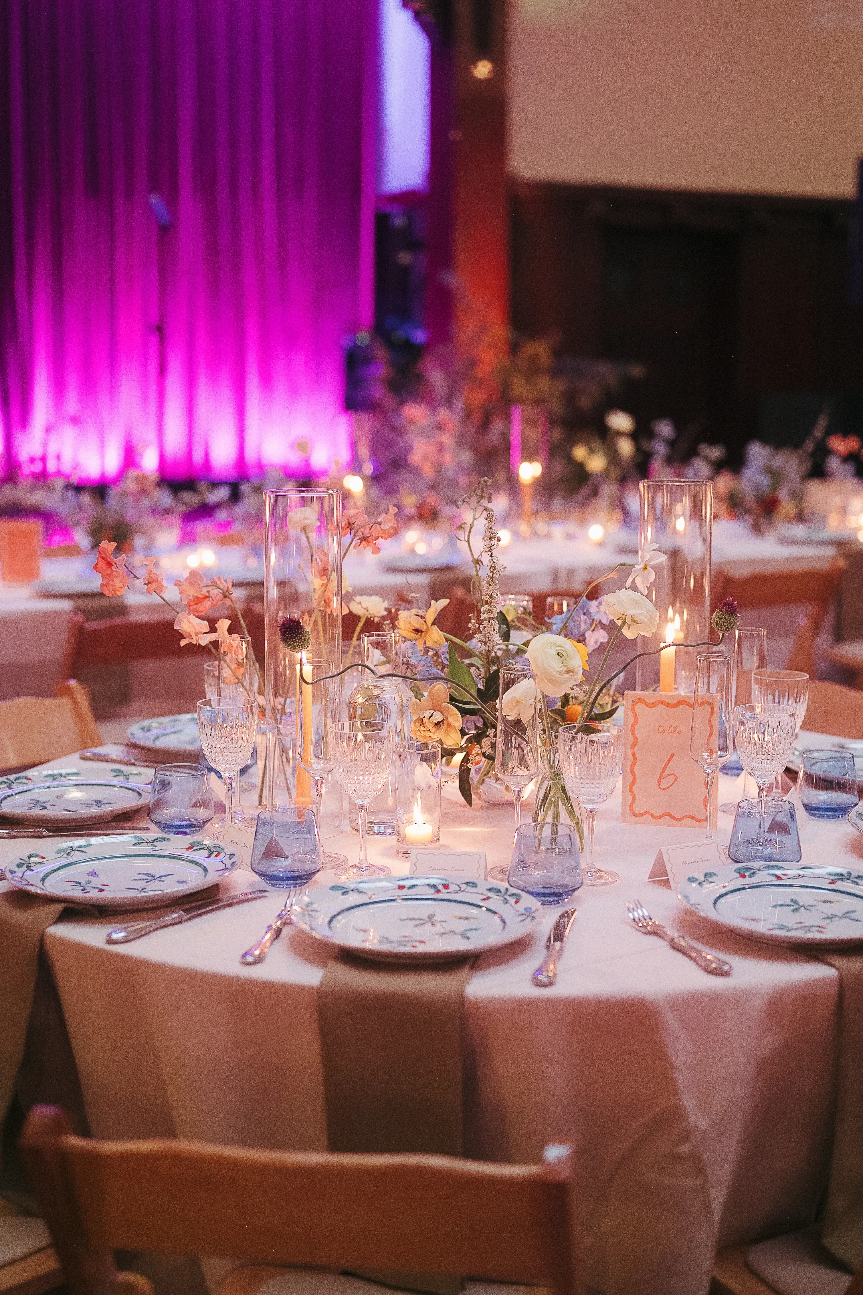 Elegant round table set for an event with floral plates, glassware, candles, and a centerpiece with flowers, under pink-purple lighting.